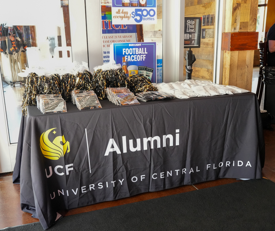 Table draped with a UCF Alumni banner, set up with pamphlets and gold/black tassels for an event, posters on the wall behind.