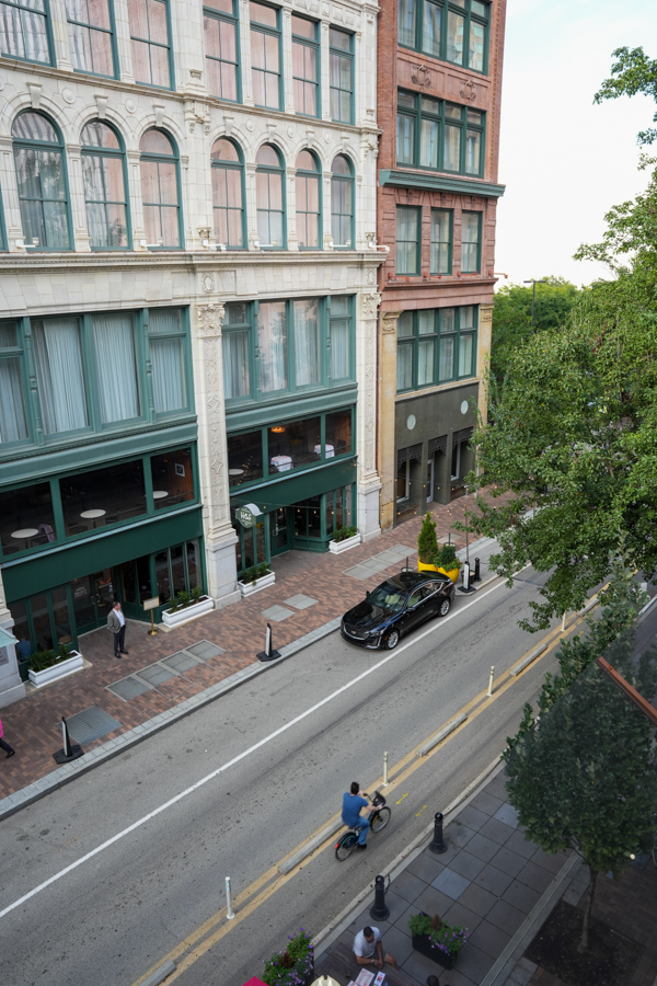 From above, a city street with an ornate white building and green storefronts, a parked black car, a cyclist, and pedestrians on the sidewalk.