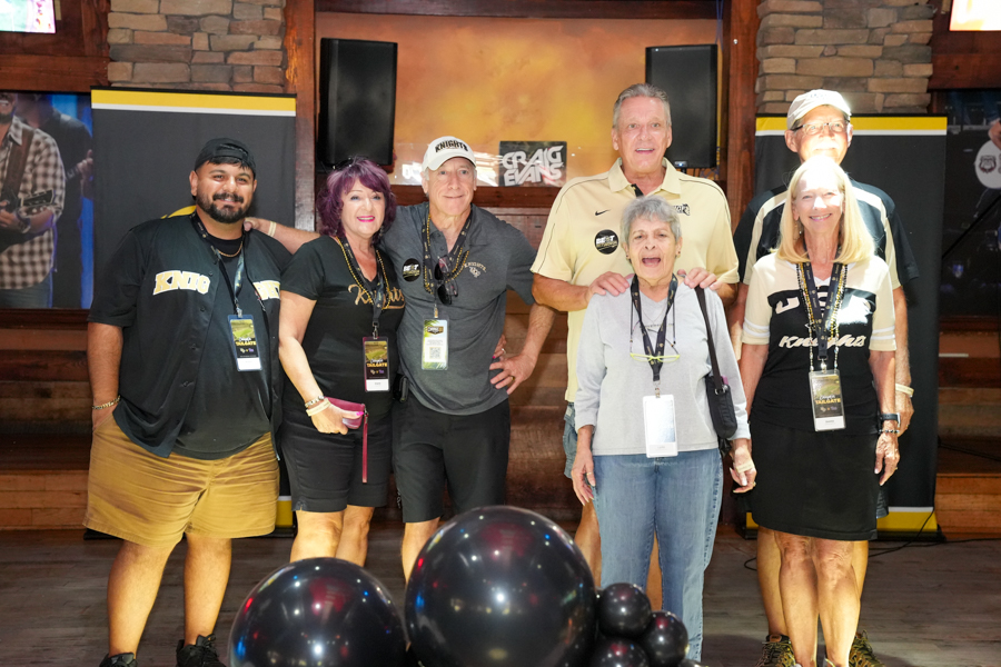 Seven people pose together for a group photo at an event, wearing lanyards and badges with a rustic stage backdrop and balloons in the foreground.