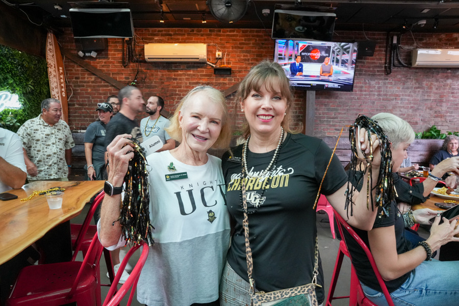 Two smiling women posing at a social event, each holding black and gold bead tassels in front of a brick-wall bar setting.