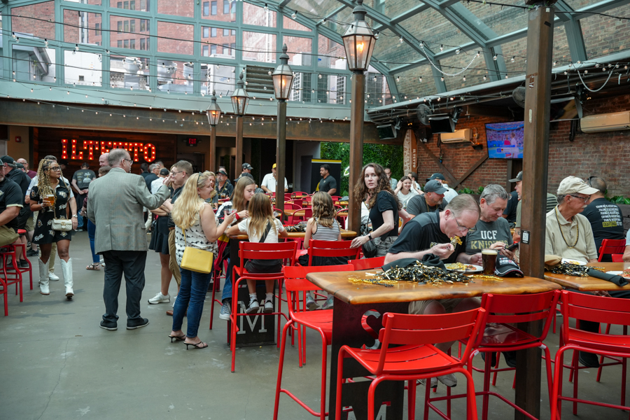 People mingle at a casual indoor courtyard with red chairs and string lights overhead.