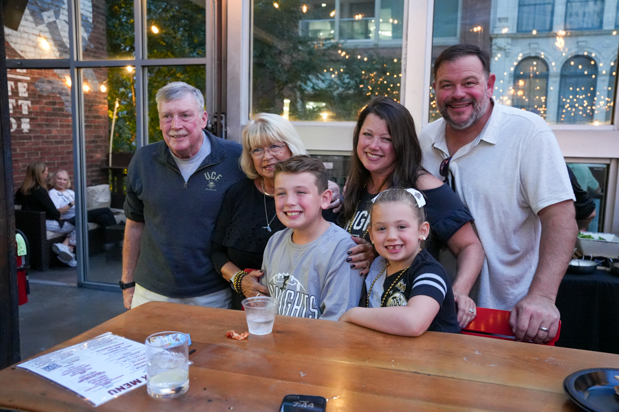 Six people of various ages posing and smiling around a wooden table at an outdoor patio with string lights in the background.