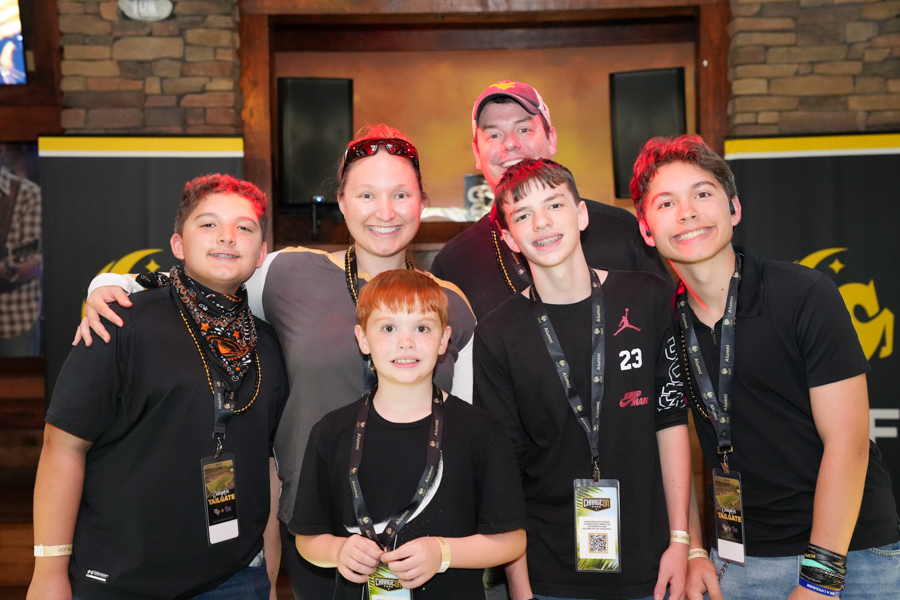 Six people posing together at an event, all smiling and wearing lanyards around their necks in a venue with brick walls and dark banners behind them.