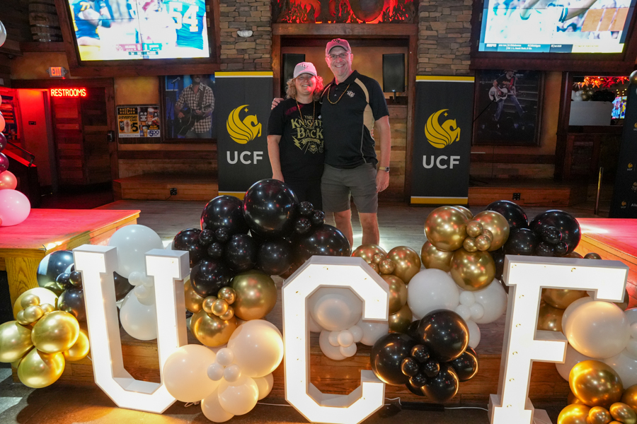 Two people posing for a photo at a UCF event in a bar, with a balloon arch and large illuminated letters in front (LUCF).