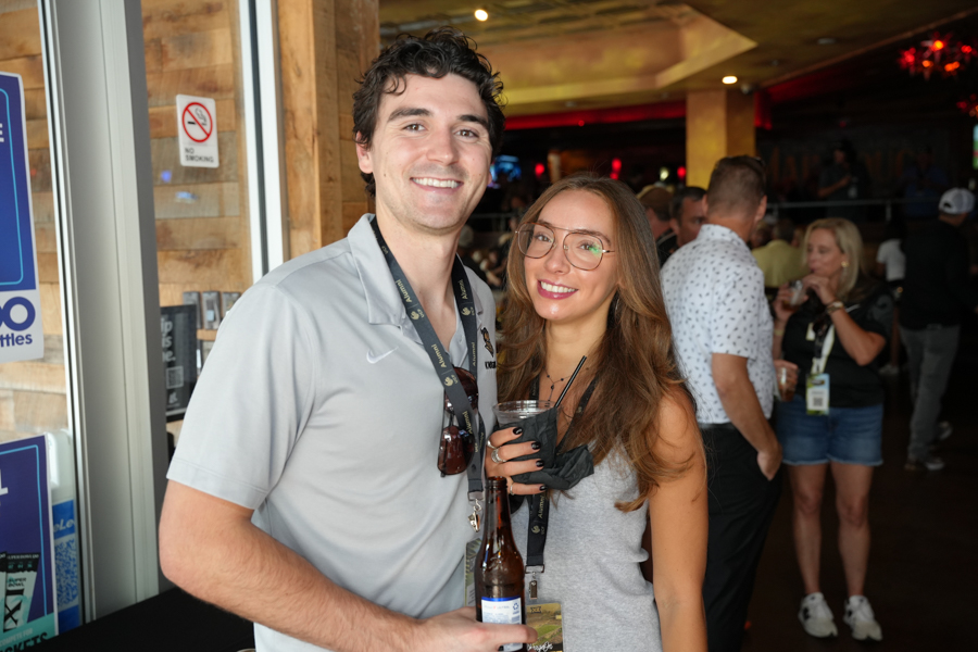 Smiling man and woman pose with drinks at a crowded indoor event, both wearing lanyards and badges.