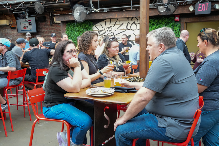 Group of people seated at a wooden table with drinks and plates, chatting in a casual indoor-outdoor bar setting with red chairs behind them