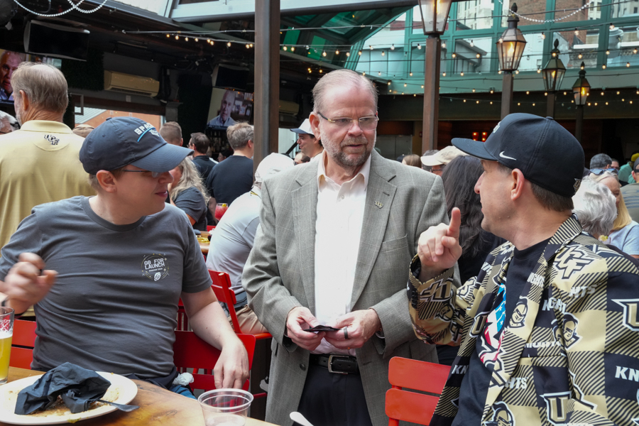 Three men mingle at an outdoor cafe; a man in a gray suit speaks with a man in a patterned jacket, while another man eats at the table nearby.