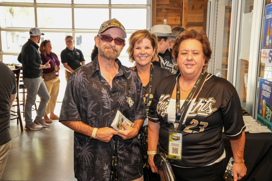 Three adults in the foreground at an indoor event pose for a photo, wearing lanyards and smiling at the camera (two women, one man).