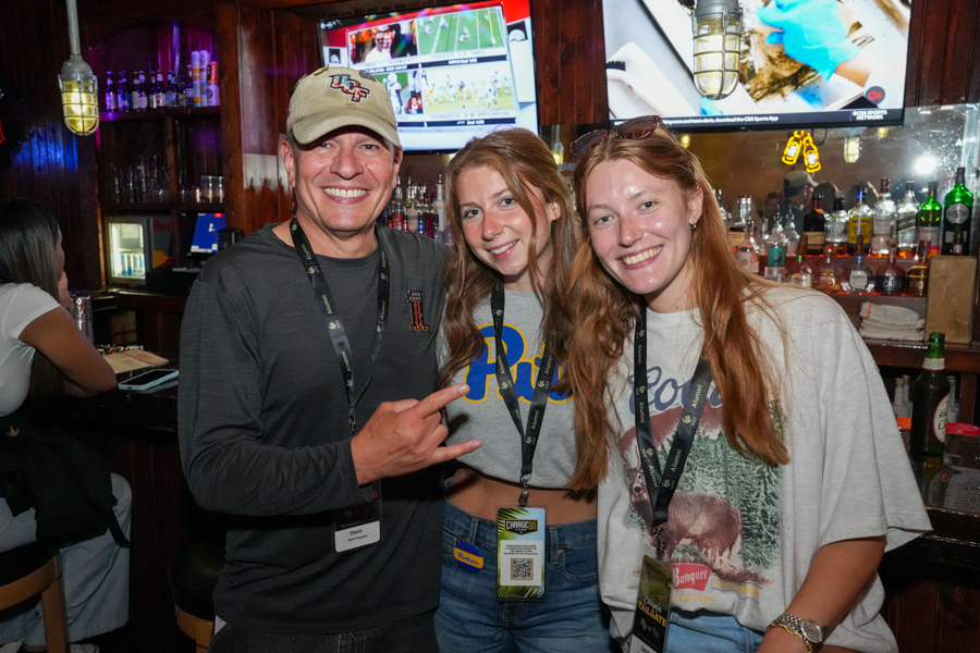 Three smiling people pose together at a bar, wearing lanyards and casual shirts without focusing on appearances in detail.]