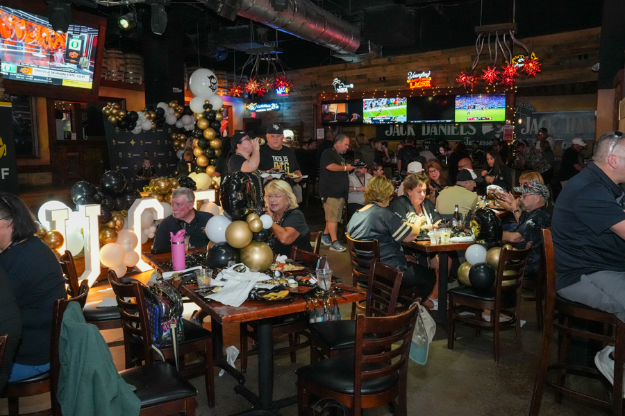 Festive restaurant scene with gold and black balloons and illuminated decor; groups of people dine and chat at wooden tables while TV screens show sports in the background.