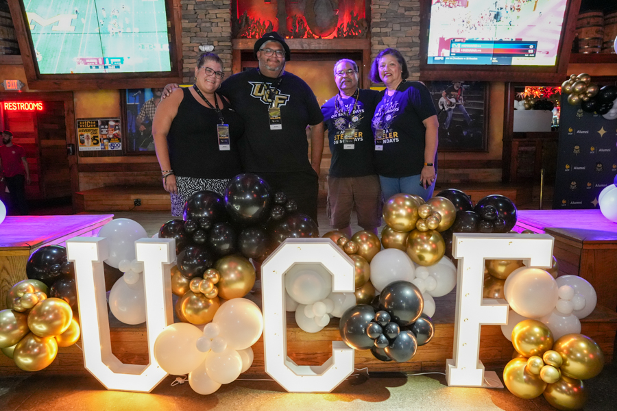 Four people wearing lanyards pose behind large illuminated UCF letters on a stage, surrounded by black, white, and gold balloons.",