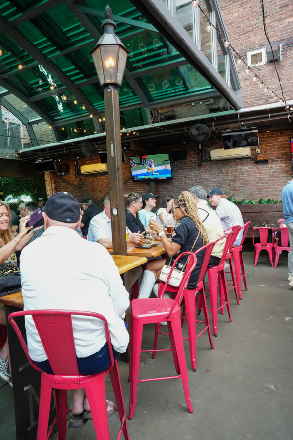 Group of people seated at a long wooden table on an outdoor patio with pink metal chairs and string lights.