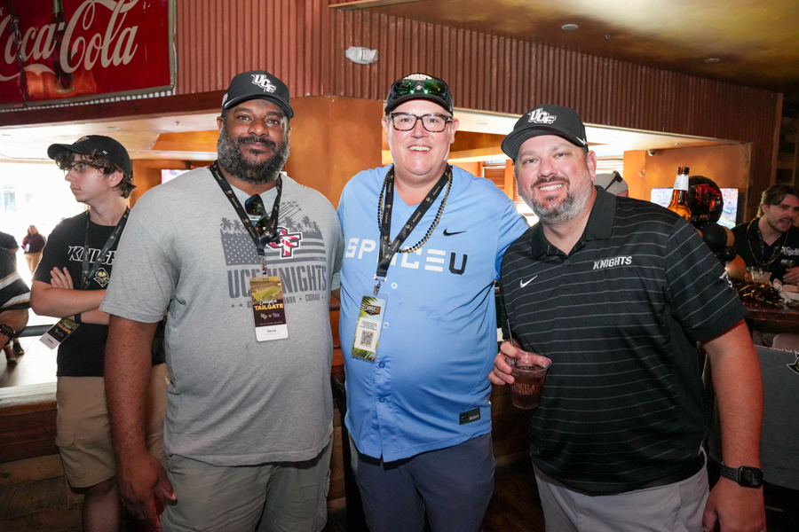 Three men wearing event lanyards pose together in a bar setting, front and center smiling for the photo with a Coca‑Cola sign in the background, two wearing Knights apparel and badges.