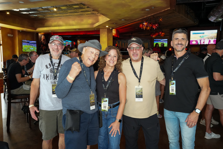 Group of five smiling adults posing for a photo at a bar or event venue, wearing lanyards and casual clothing.