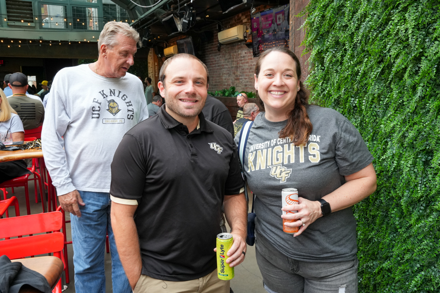 Two smiling friends pose for a photo at an outdoor venue; the woman wears a University of Central Florida Knights t‑shirt and holds a drink, the man holds a yellow can.