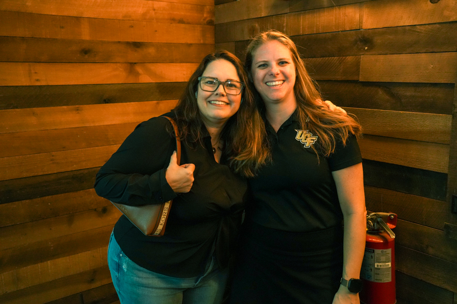 Two women smiling for a photo indoors against a wood-paneled wall; one gives a thumbs‑up, the other wears a black shirt with a UCF logo.