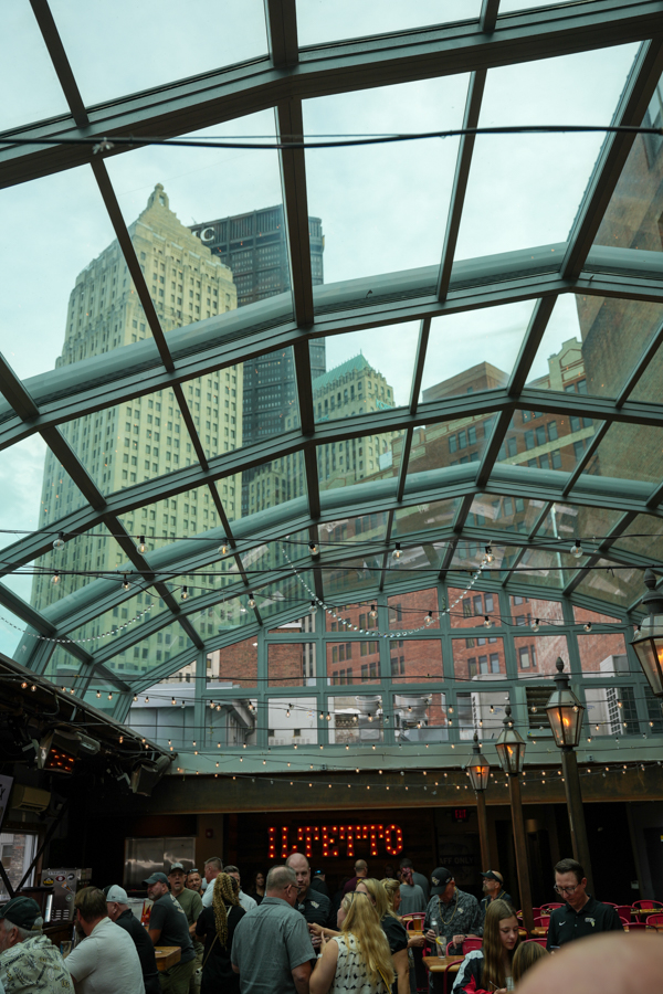Crowded indoor market/restaurant under a curved glass atrium with string lights and diners at tables; city buildings visible outside the glass ceiling.