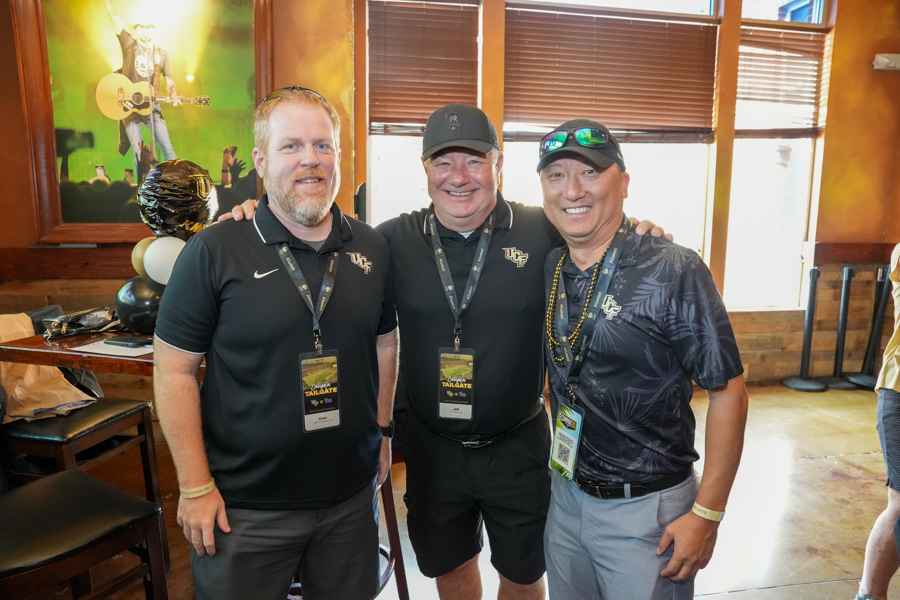 Three smiling men stand together with arms around each other at a UCF tailgate event, wearing lanyards and badges in a warm indoor space.