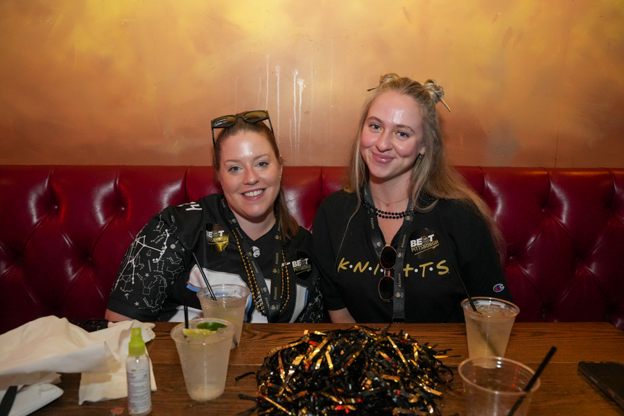 Two women sit in a red tufted booth, smiling at the camera with drinks on the table and festive confetti in front of them in a warm, casual bar setting.