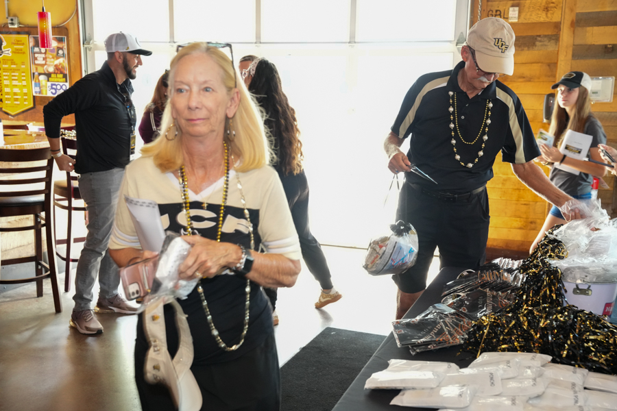 Older woman in the foreground wearing a white and black shirt with a beaded necklace, holding papers near a table at a busy indoor event.