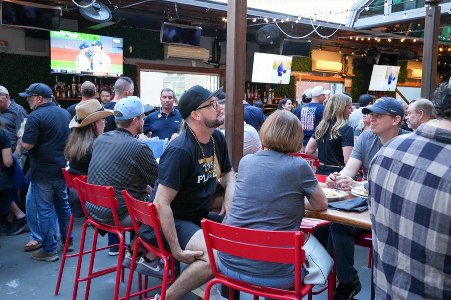 Crowded outdoor bar with people sitting on red chairs at tables, watching a baseball game on multiple TVs.