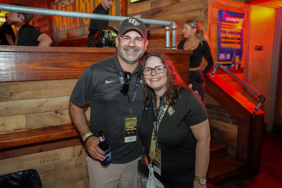 Smiling man and woman pose together at a UF tailgate event, wearing lanyards; the man holds a beer bottle in a wooden-bar venue.