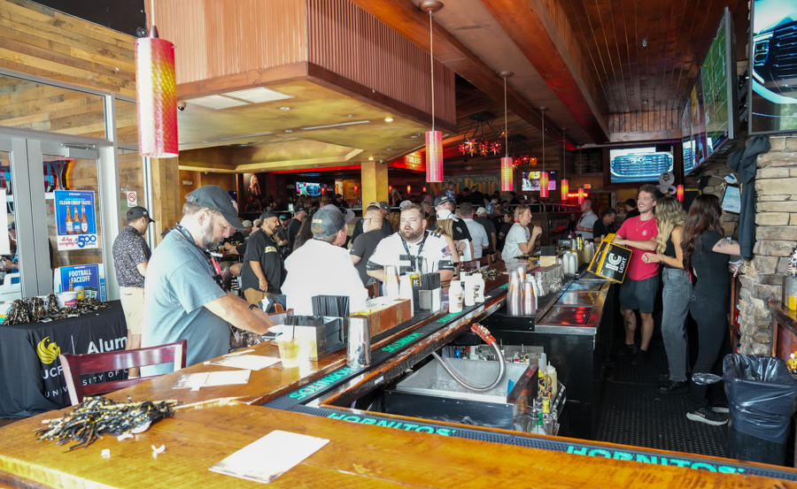 Crowded bar with bartenders serving drinks along a long wooden counter as patrons chat and wait for orders.