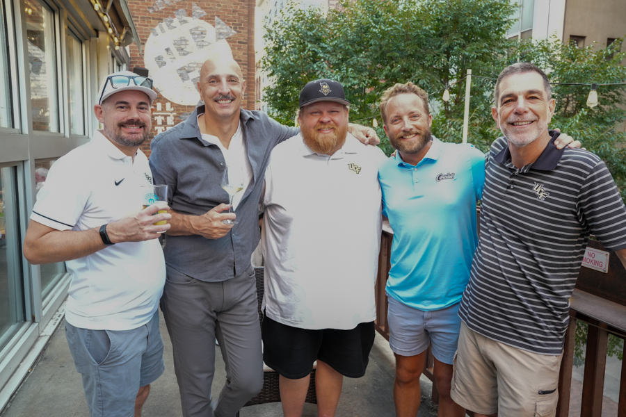 Five men smiling and posing together on a balcony, each holding a drink and with their arms around each other.