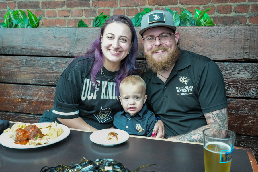 Family of three at a restaurant booth: mother with purple hair, father with beard and glasses, and a toddler in UF polo smiles at the camera with plates of food in front.