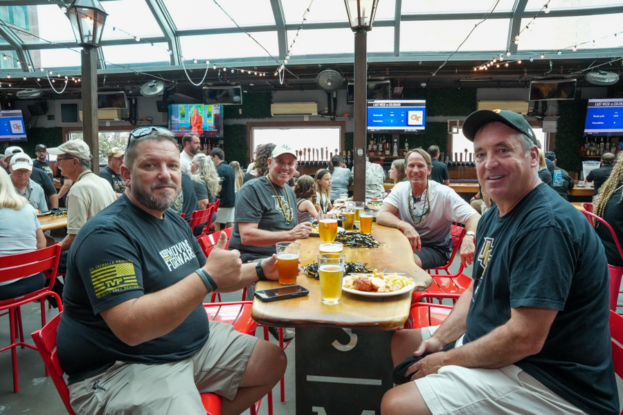 Group of people at a long wooden table in a busy bar, smiling with beers and plates of food.