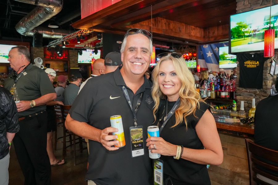 Smiling man and woman pose together at a bar, each holding a canned drink and wearing event lanyards.
