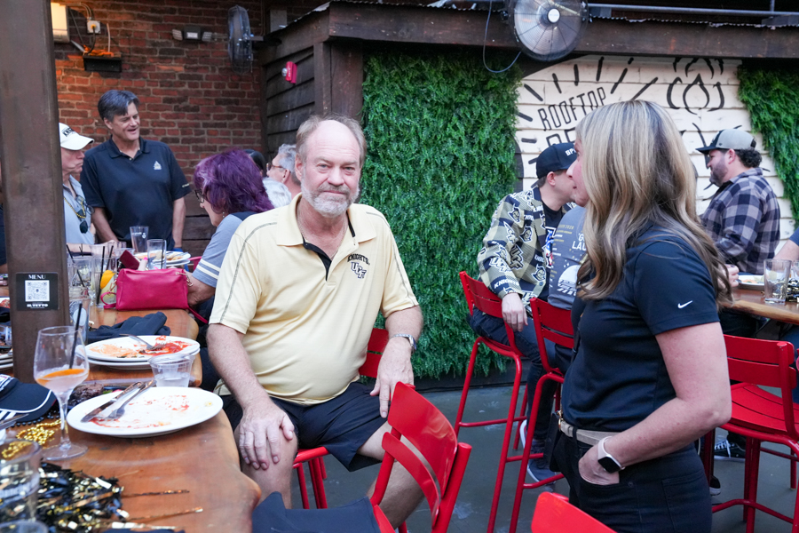 Outdoor rooftop dining scene with people chatting at a long wooden table; a man in a yellow polo sits near plates while a woman in black talks nearby, red stools around them.