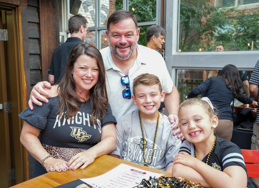 Family of four posing for a photo at an outdoor event; mom wears a Knights shirt, dad stands behind with sunglasses, kids sit at the table.
