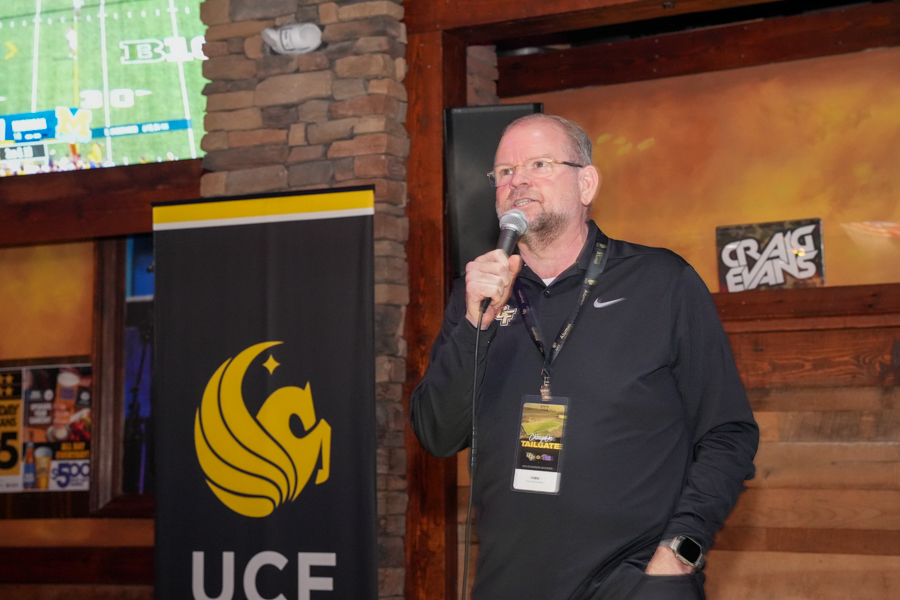 Man in a black polo with a lanyard speaks into a handheld microphone at a tailgate event, with a UCF banner nearby and a stone/wood backdrop.
