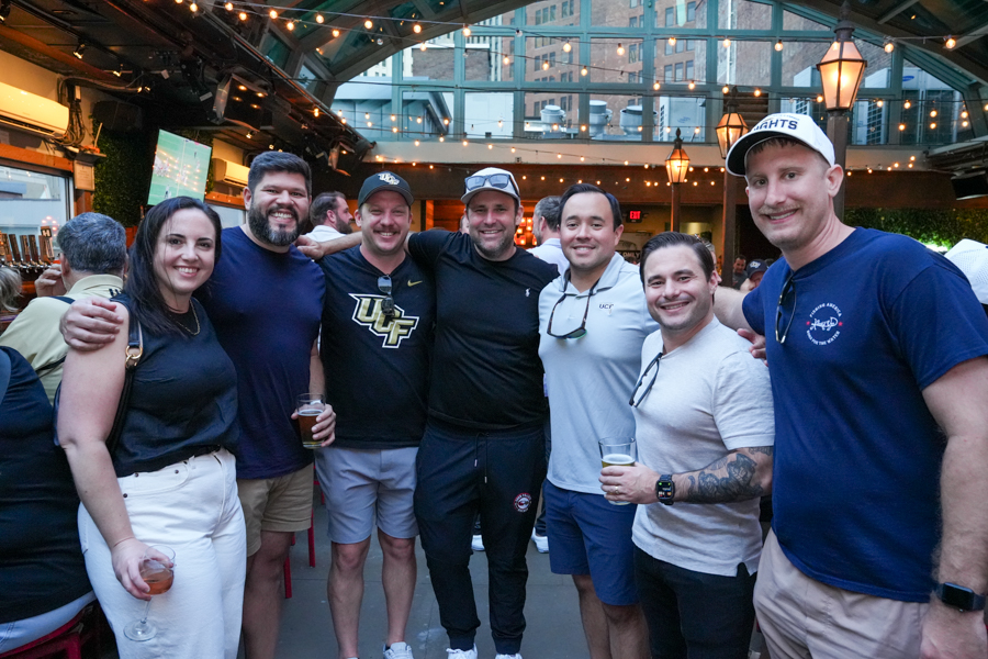Group of friends posing together at a casual indoor-outdoor event, holding drinks under string lights.