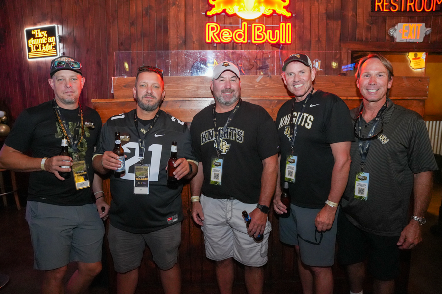 Five men stand shoulder to shoulder in a bar, each holding a beer and wearing lanyards; neon Red Bull sign glows behind them.