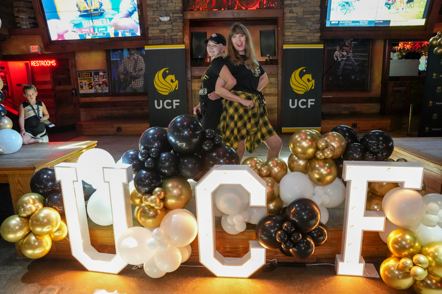 Two adults posing on a stage with large illuminated UCF letters and gold, black, and white balloons surrounding them.
