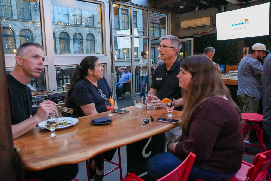 Group of adults seated around a curved wooden bar/table indoors with city buildings visible through large windows, chatting and sipping drinks.