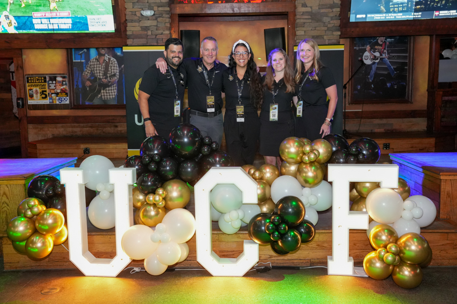 Five staff members posing on a stage behind a balloon display and large illuminated UCF letters, at an event.