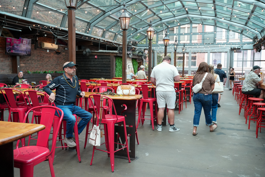 Indoor patio/bar area with red metal chairs and wooden tables under a glass roof; a man in a cap sits at a high table while others stand or walk by.