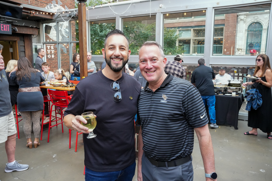 Two men smiling and posing for a photo at an outdoor gathering, one holding a glass of white wine.