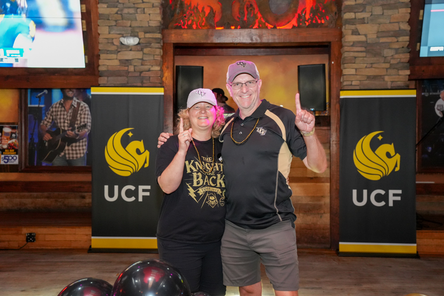 Two smiling adults pose for a photo at a UCF event, raising a finger each, with UCF banners in the background.