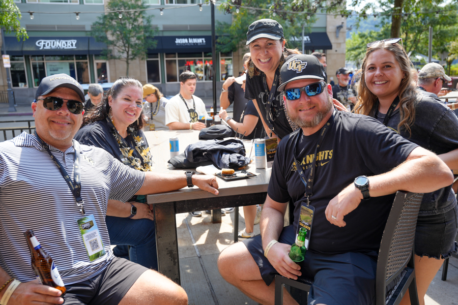 Group of five friends at an outdoor cafe table, smiling for the camera; two men hold beers, others lean in from behind in a sunny urban setting