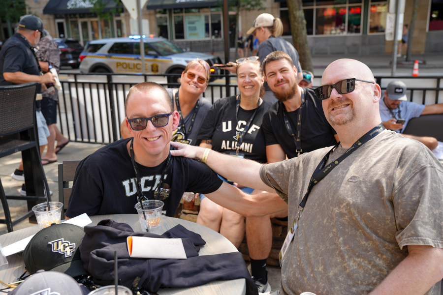 Group of smiling UCF Alumni posing at an outdoor event, with a man in sunglasses at the foreground and others behind him at a sidewalk gathering.