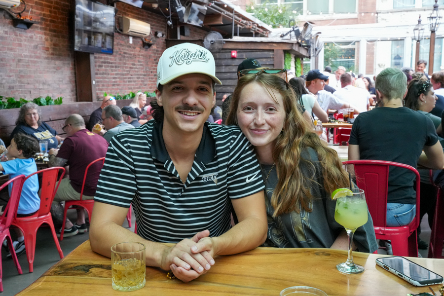 Couple sitting at a wooden table at a busy outdoor restaurant, smiling with drinks in front.