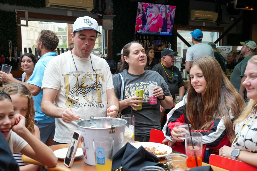 Group of friends at a busy bar, sharing pizza and drinks around a table while chatting and laughing.