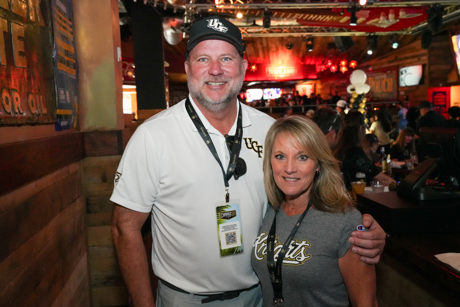 Man wearing a white UCF polo and cap poses with a smiling woman in a gray T‑shirt at a crowded bar event.