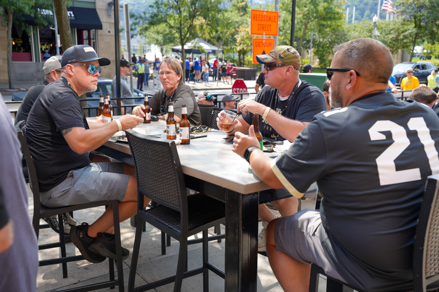 Four men sit at a high outdoor table, drinking bottled beer on a sunny street sidewalk.
