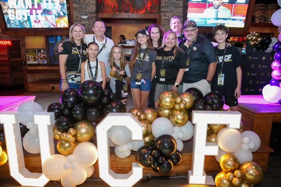 Group of people posing behind illuminated UCF letters adorned with black, white, and gold balloons at an indoor event venue.