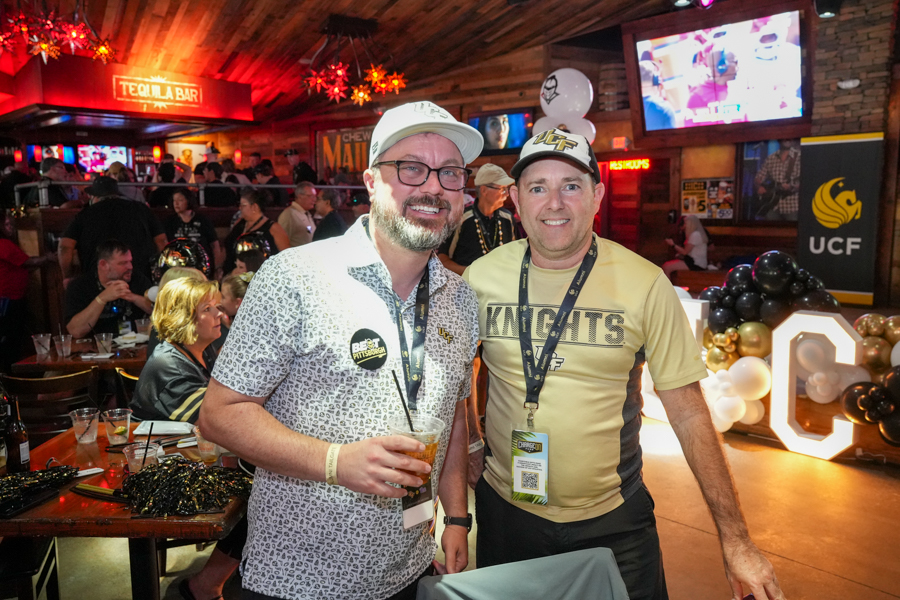 Two men pose for a photo at a crowded bar/event, one in a UCF Knights shirt and cap, the other in a patterned polo, both wearing lanyards and badges with drinks in hand.